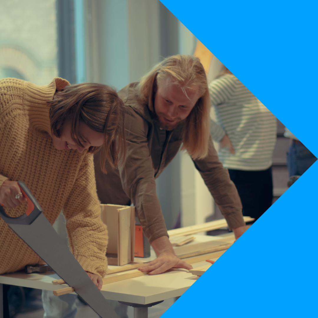 Kuvassa on kaksi henkilöä ja toinen heistä sahaa. Oikeassa ja vasemmassa nurkassa on kaksi kolmio-elementtiä.